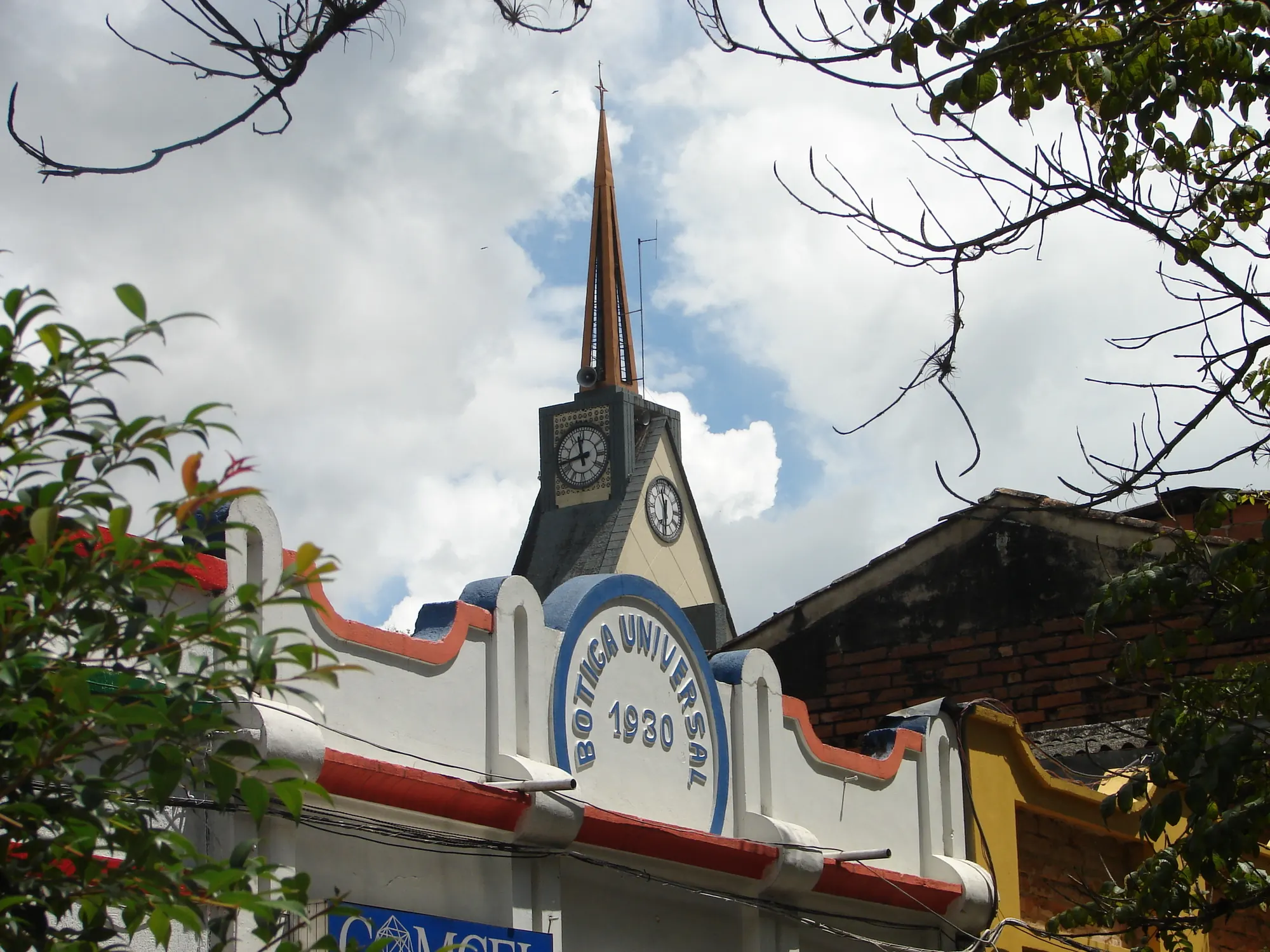 Fachada de la «Botica Universal 1930», con remates curvos pintados y la torre del reloj de la iglesia detrás.