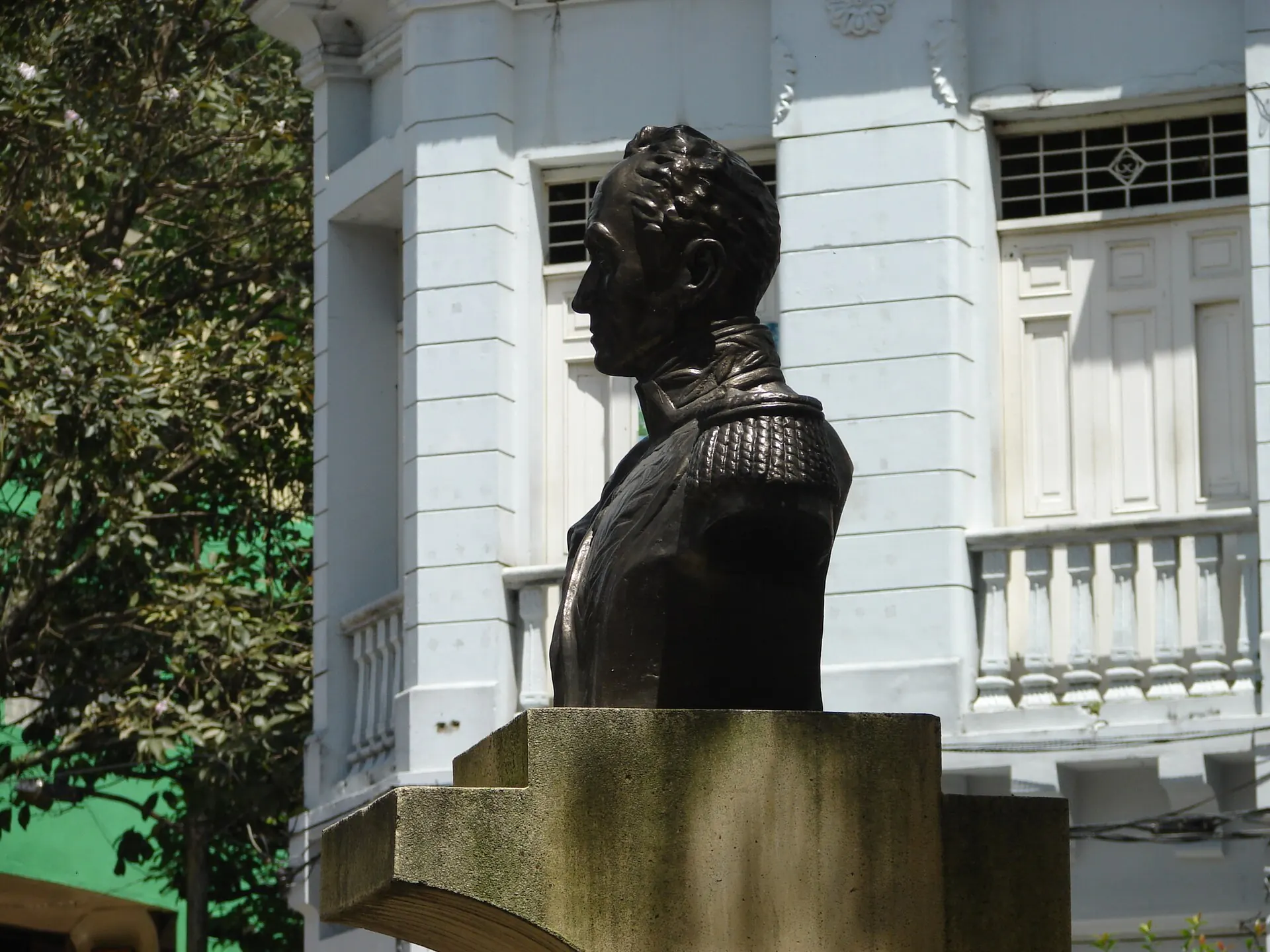 Busto de Simón Bolívar en bronce sobre pedestal, con fachada blanca de balcones al fondo.