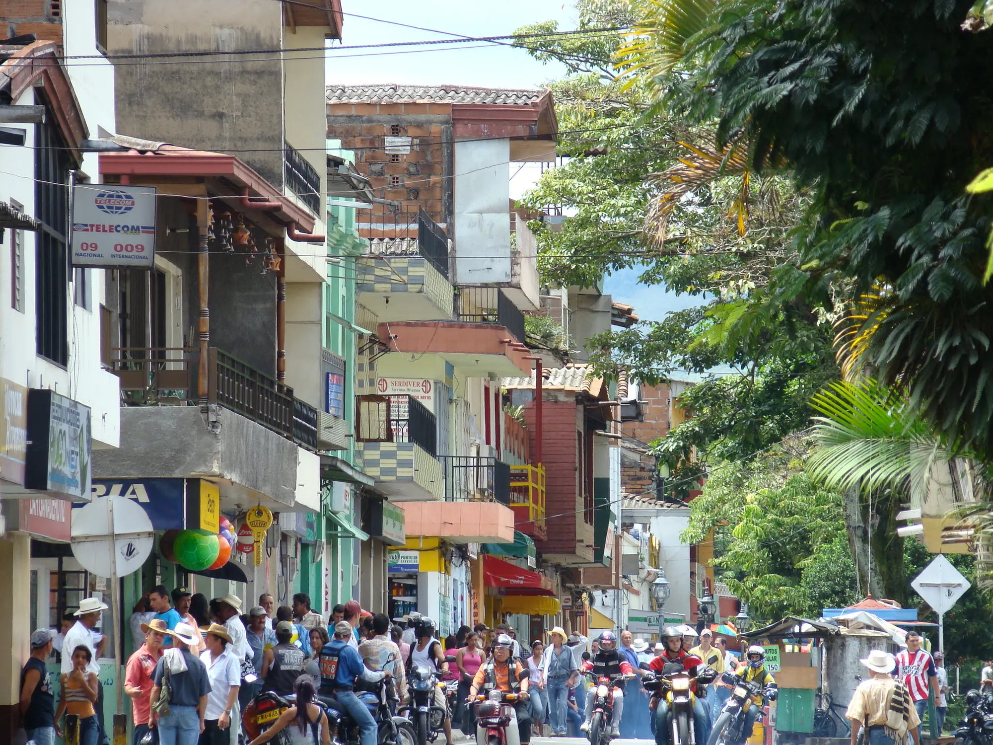 Calle comercial llena de gente y motos, con fachadas coloridas y montaña al fondo.