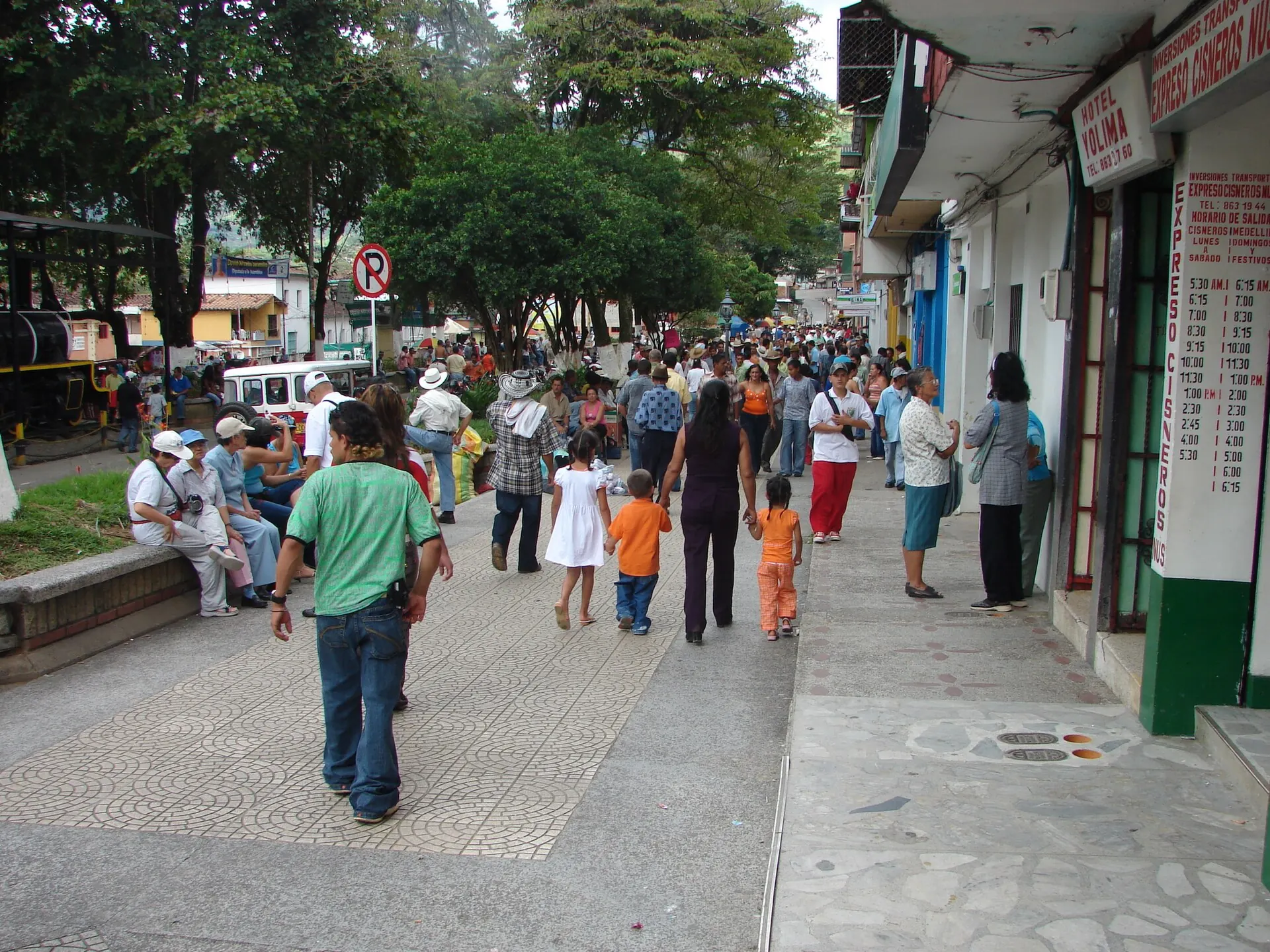 Calle principal peatonal con mucha gente un domingo, comercios a la derecha y árboles del parque a la izquierda.