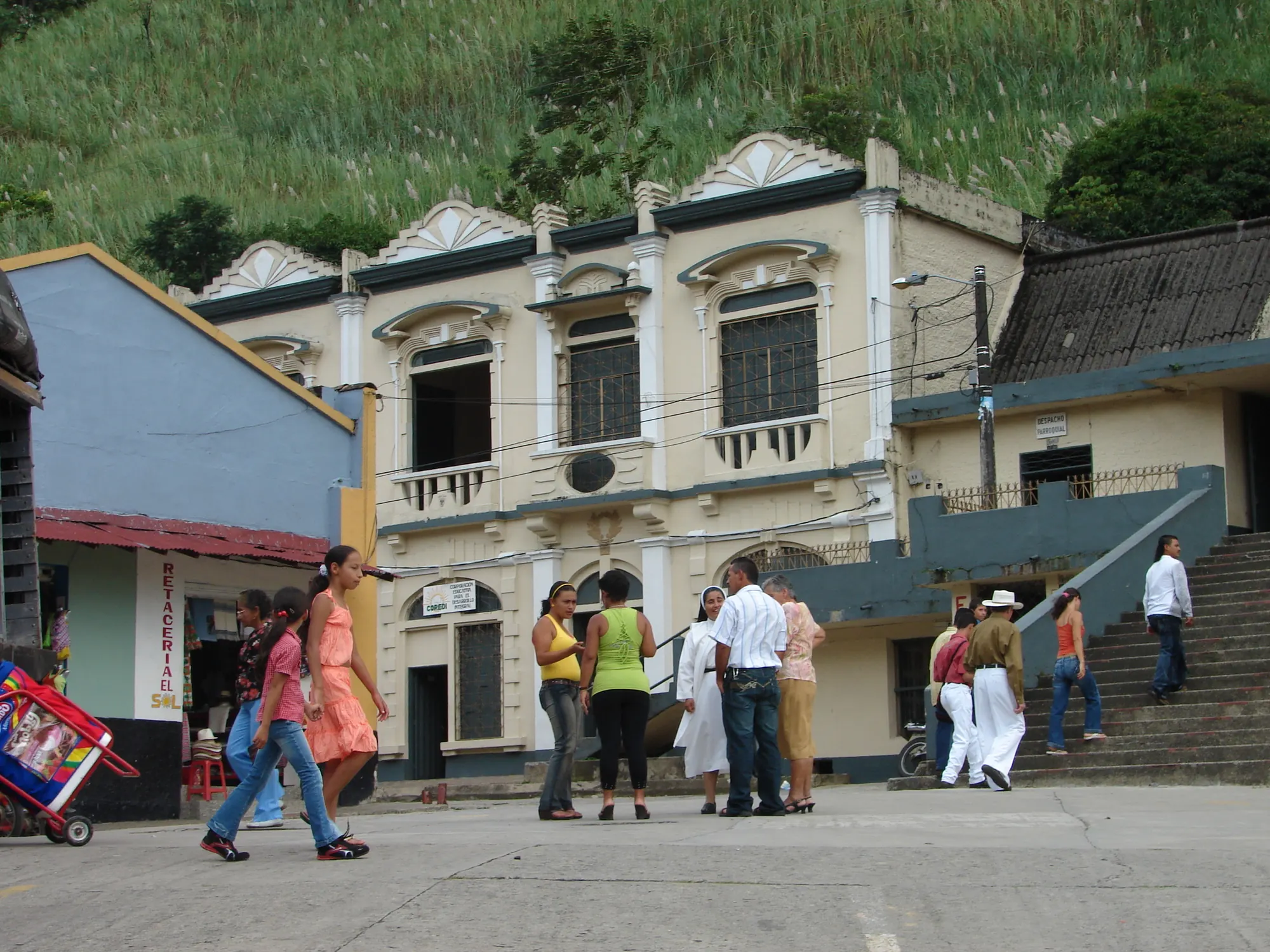 Edificio de dos plantas con balcones y remates ornamentales, escalinata lateral y gente conversando un domingo.