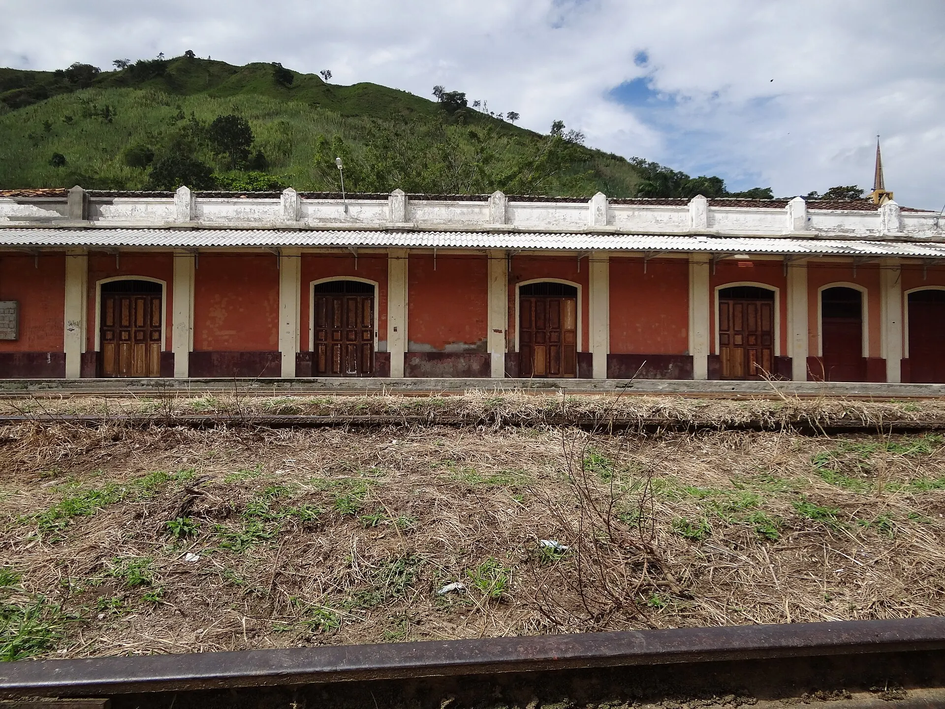 Fachada frontal de la estación vista desde las vías, con montaña al fondo.