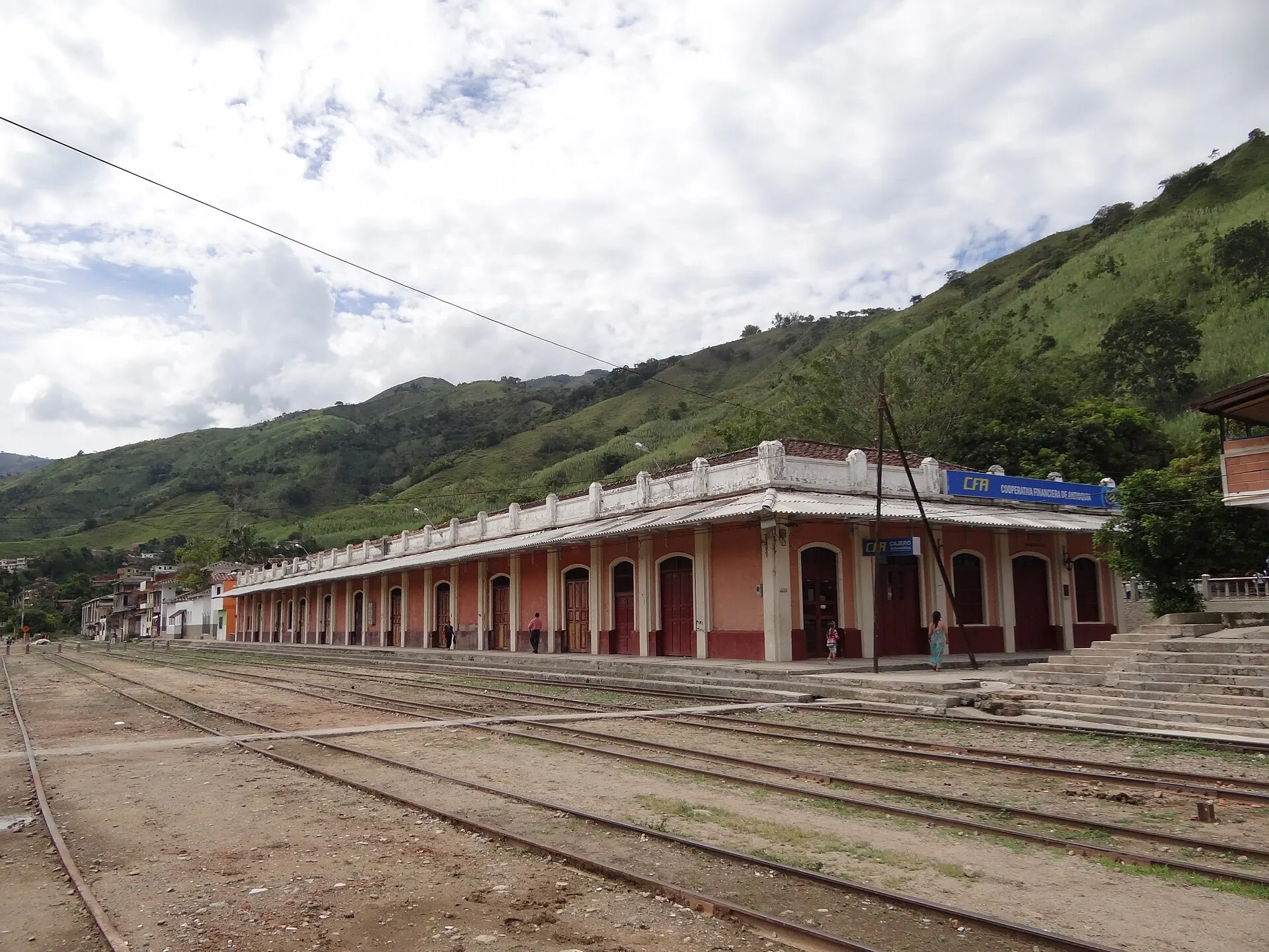 La estación en perspectiva con el patio de vías y el andén, montañas y nubes.