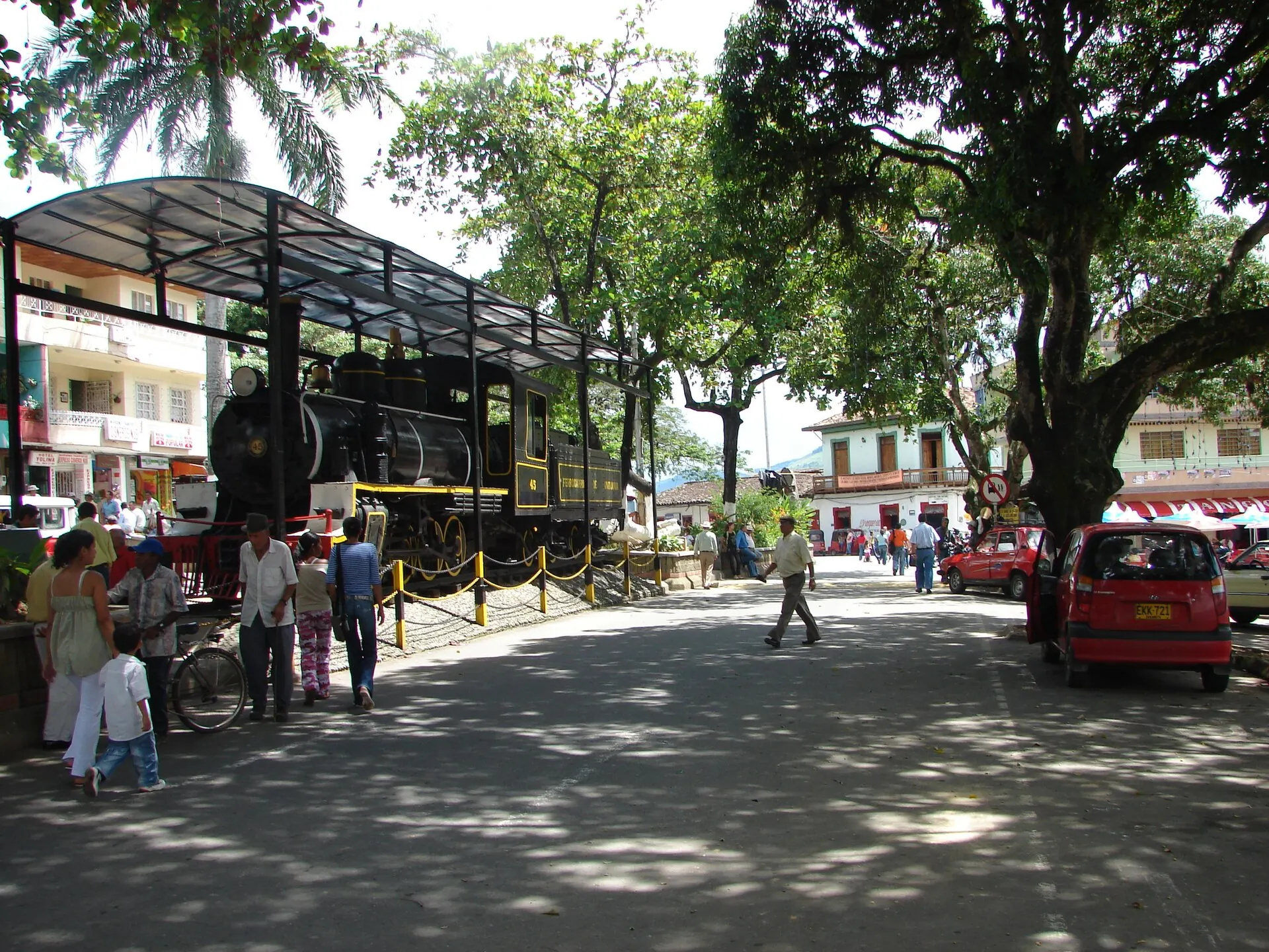 Locomotora bajo techo junto al parque, con transeúntes, árboles grandes y carros.