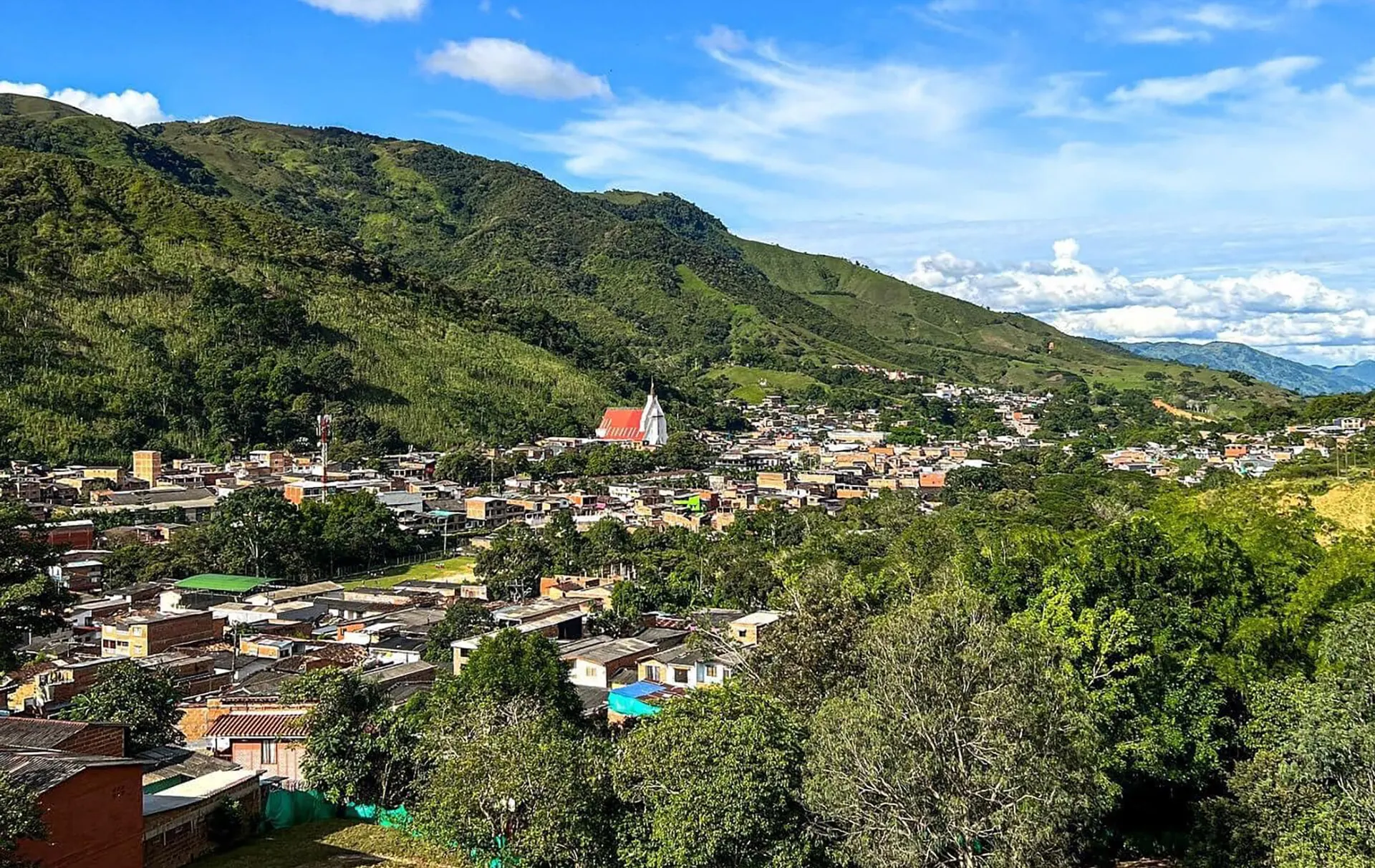 Vista panorámica del casco urbano de Cisneros en el fondo del cañón del río Nus, rodeado de montañas verdes, con cielo azul y nubes.
