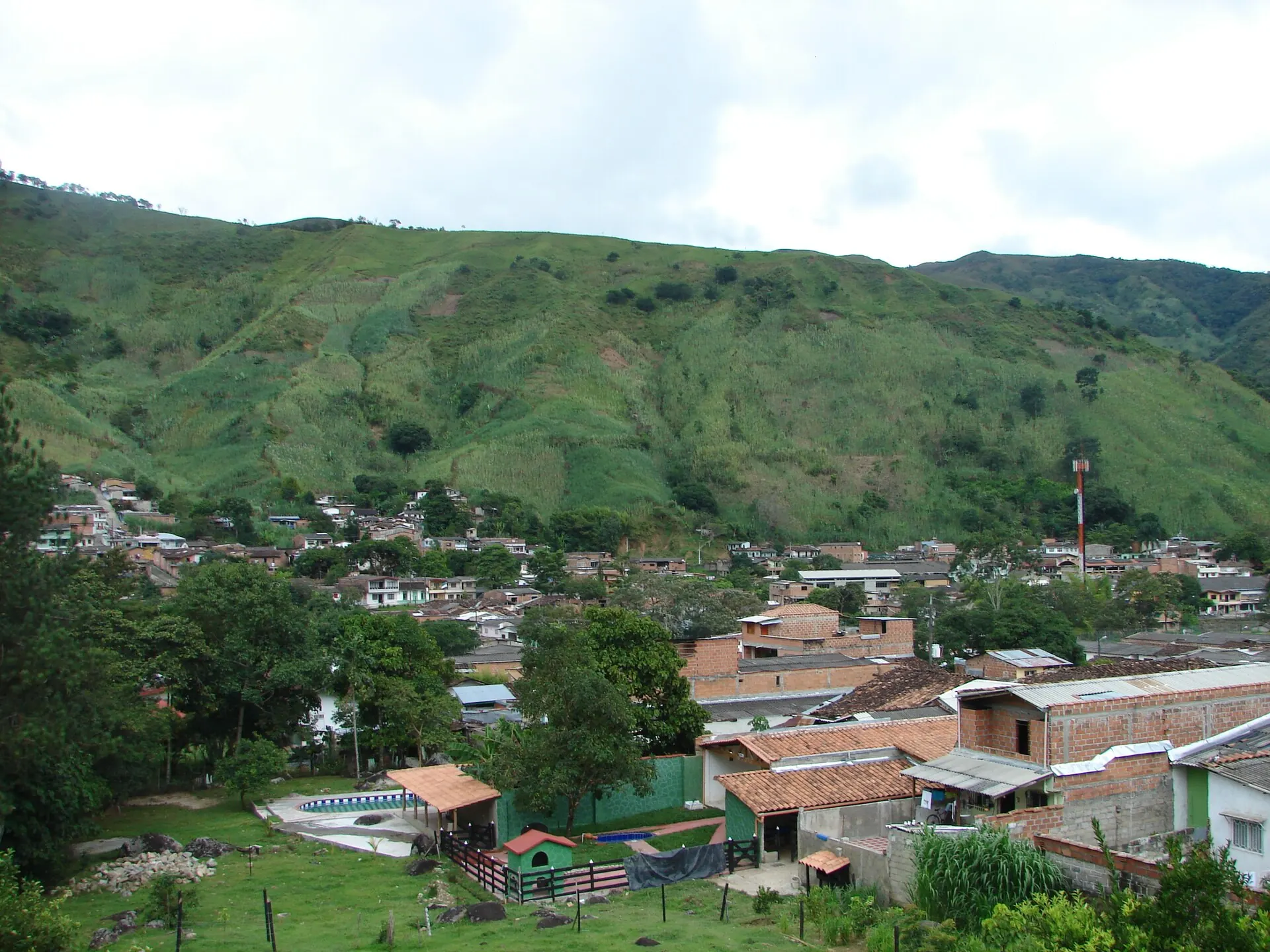 Panorámica del pueblo desde una ladera: tejados de barro y de zinc en primer plano, montañas con potreros al fondo.