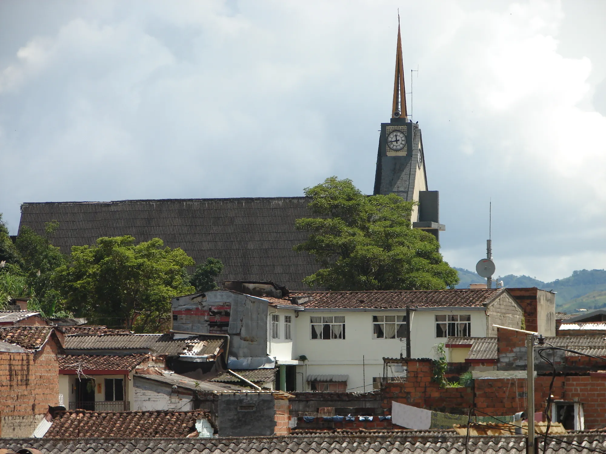 Tejados del pueblo con la torre de la iglesia (reloj y aguja) contra el cielo, árboles y montañas.