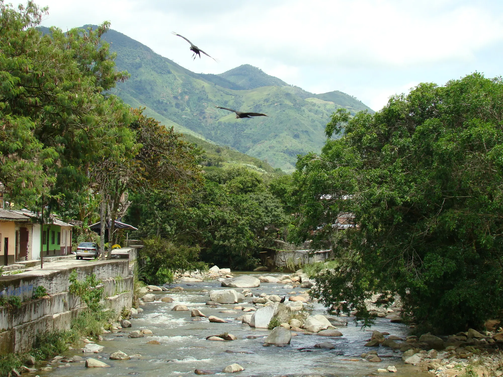 Río pedregoso canalizado junto a las casas, con gallinazos volando y montañas al fondo.