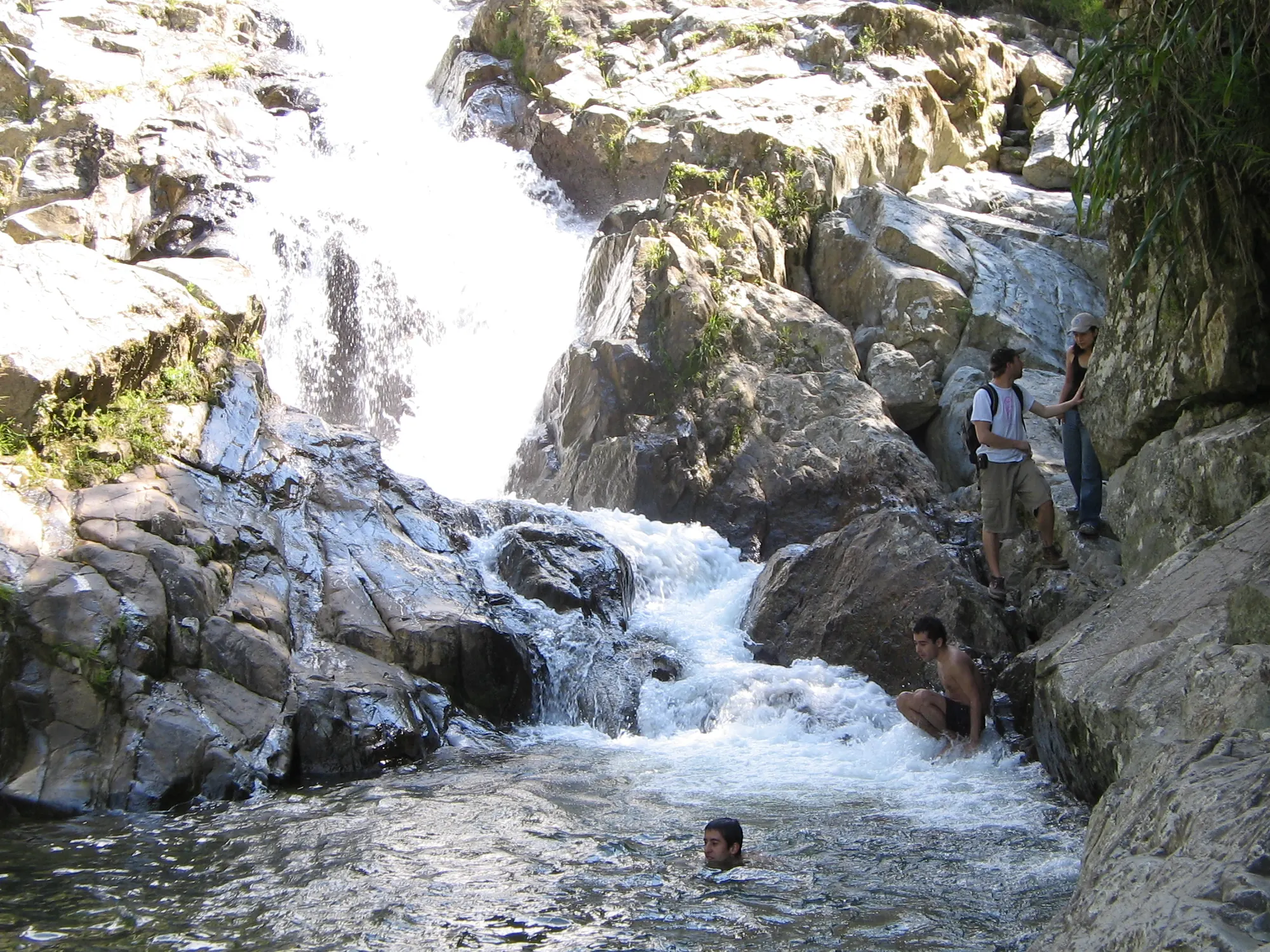 Cascada entre grandes rocas que cae a un pozo, con bañistas pequeños en el agua.