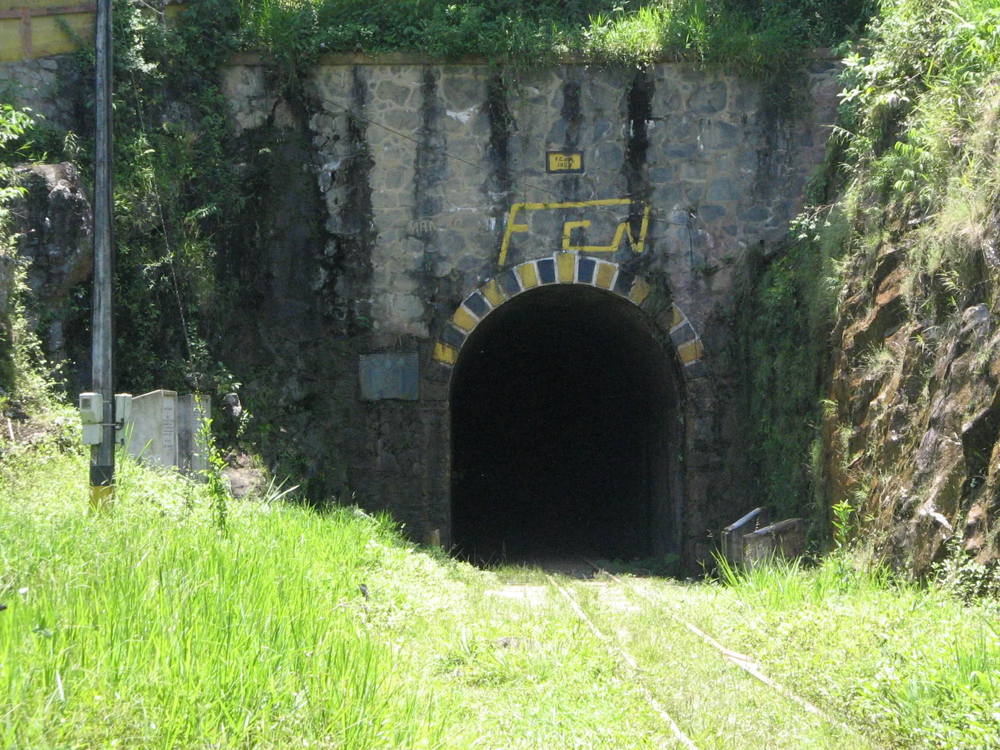 Portal occidental del túnel de La Quiebra, con su emblema amarillo, vegetación y rieles.